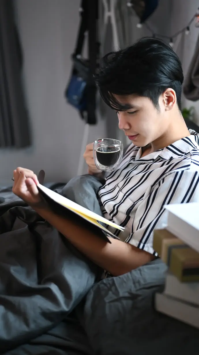 Hombre asiático tomñandose un cafe y leyendo en la cama